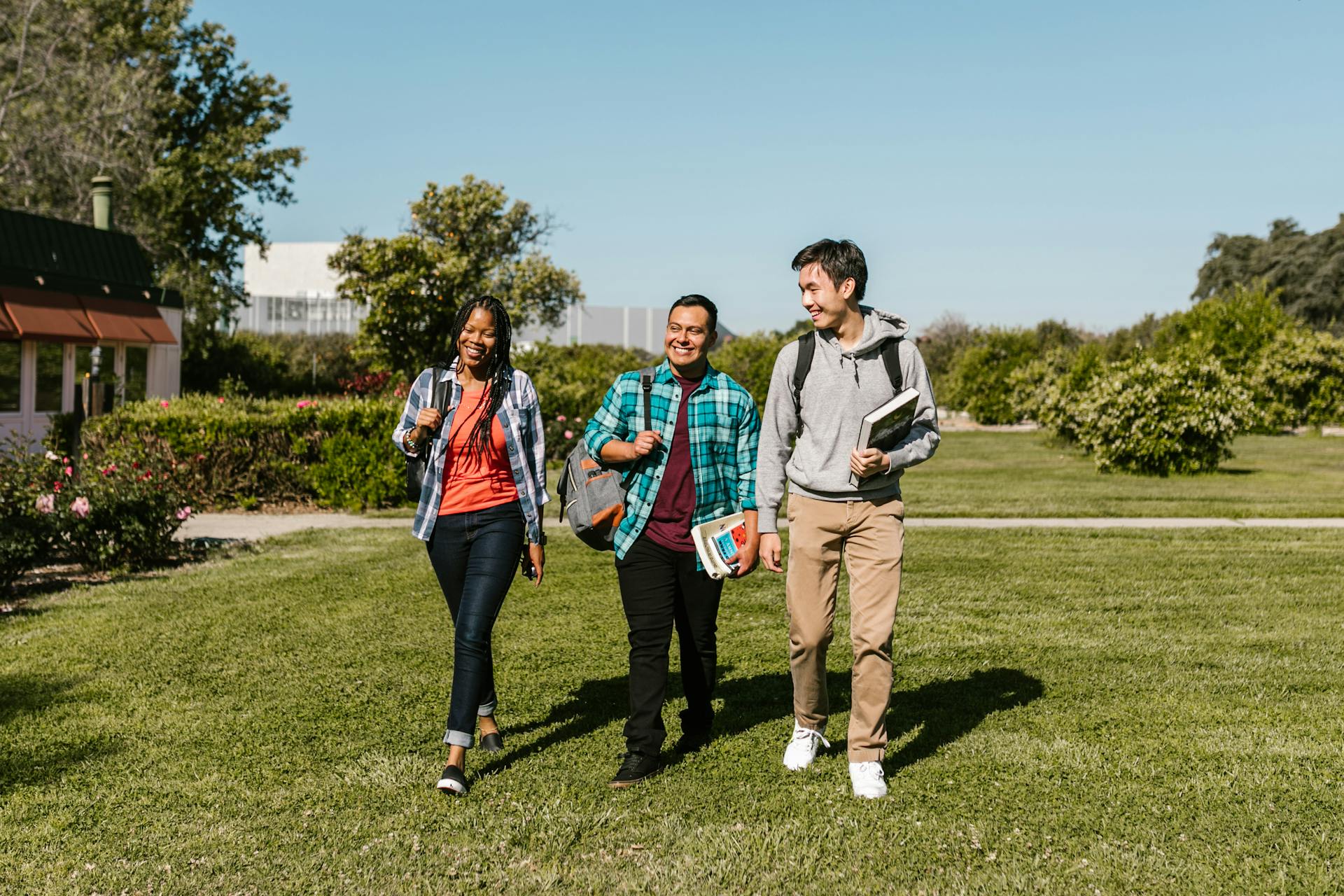 Three college students walking together on a green campus lawn, carrying backpacks and textbooks, enjoying a sunny day.