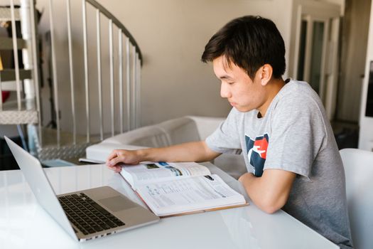 Teenage boy focuses on homework using a laptop and textbook indoors.