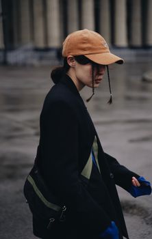Side view of unrecognizable trendy female in orange cap walking near columns of building on street on rainy autumn day