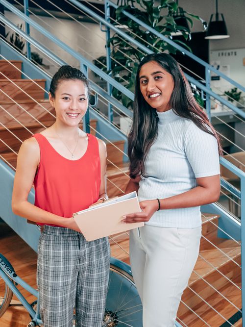 Free Two young businesswomen collaborating in modern office space, standing by stairs, holding documents. Stock Photo