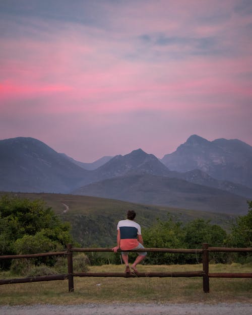 A Person Sitting on a Wooden Fence near the Mountains