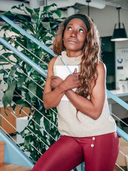 A professional young woman stands indoors, thoughtfully holding a notebook by the staircase.