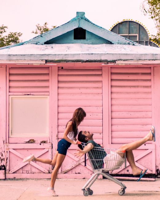 A joyful couple sharing a playful moment with a shopping cart in front of a pink building.