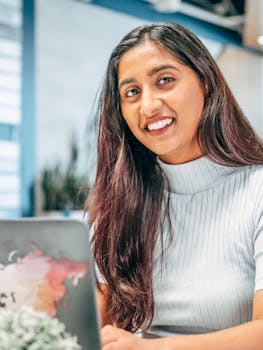 Portrait of a woman with long hair smiling at a laptop indoors, conveying positivity.