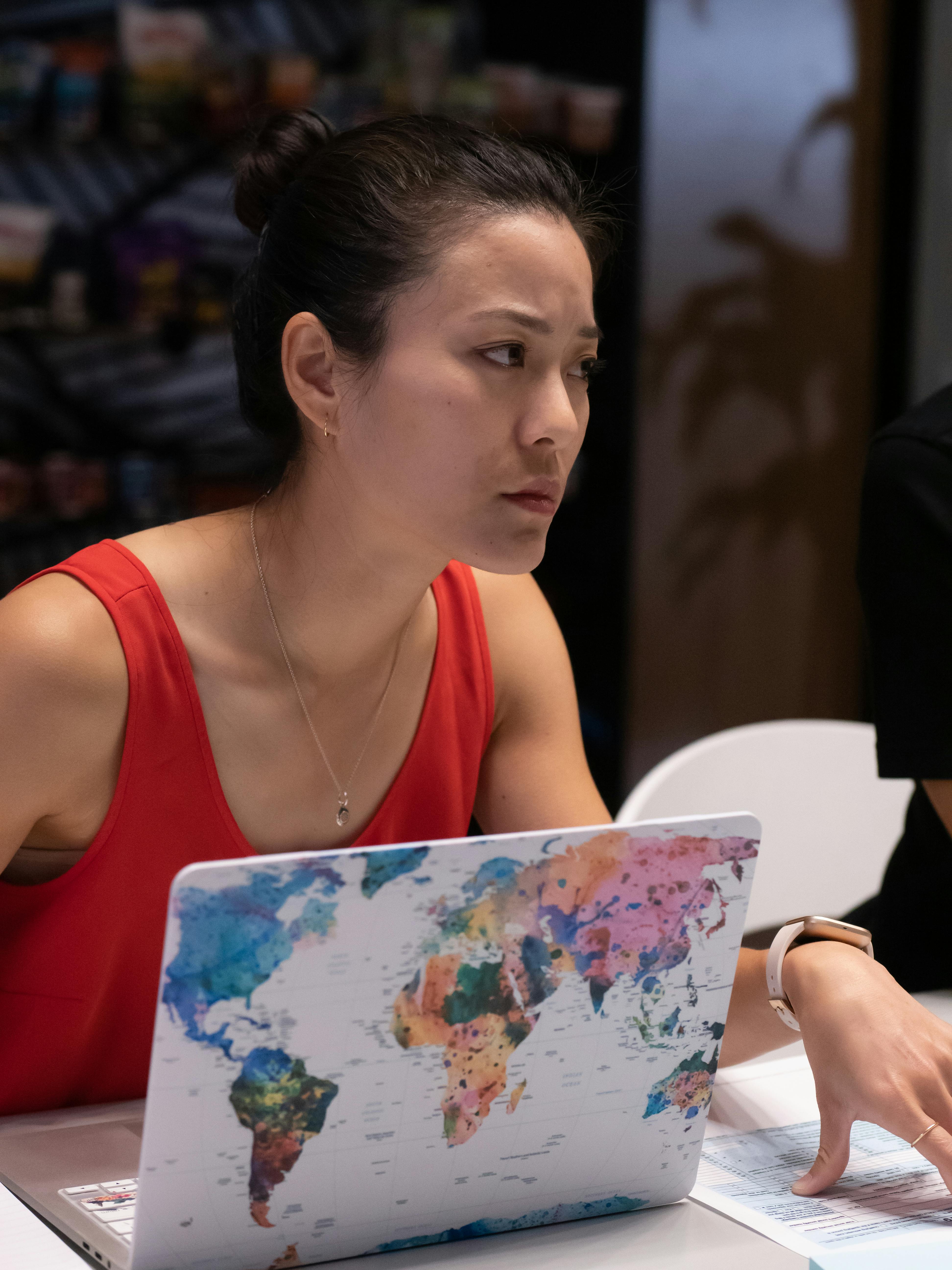 Asian businesswoman in a red tank top focused on her work at an office desk with a colorful world map laptop skin.