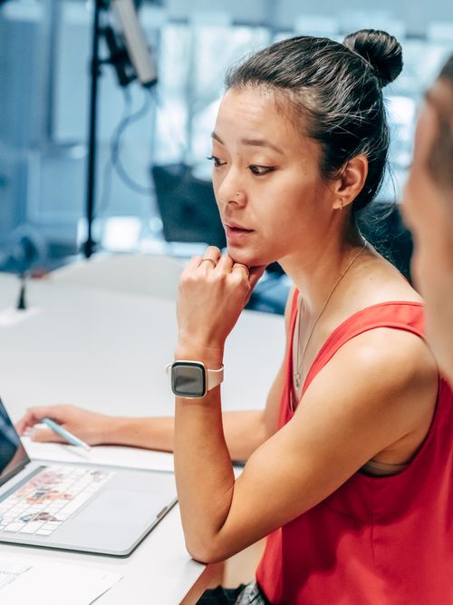 Free An Asian woman in a red top working on a laptop, appearing thoughtful in a modern office. Stock Photo