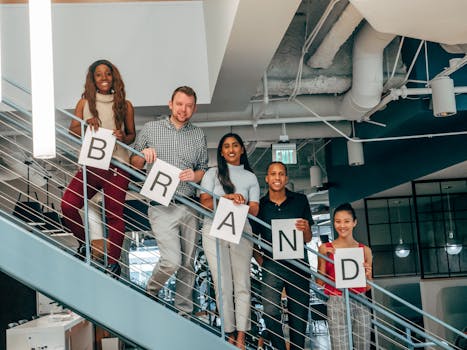 Diverse group of adults smiling and holding 'BRAND' signs on an office staircase.