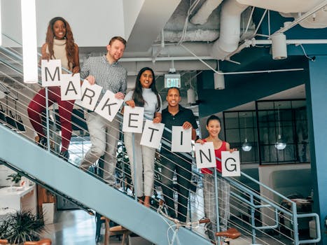 Five colleagues smiling and holding a marketing sign on an office staircase, showcasing teamwork.