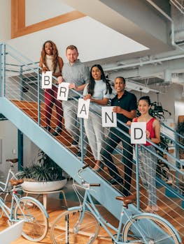 A diverse group of colleagues smiling and holding a 'Brand' sign on indoor office stairs.