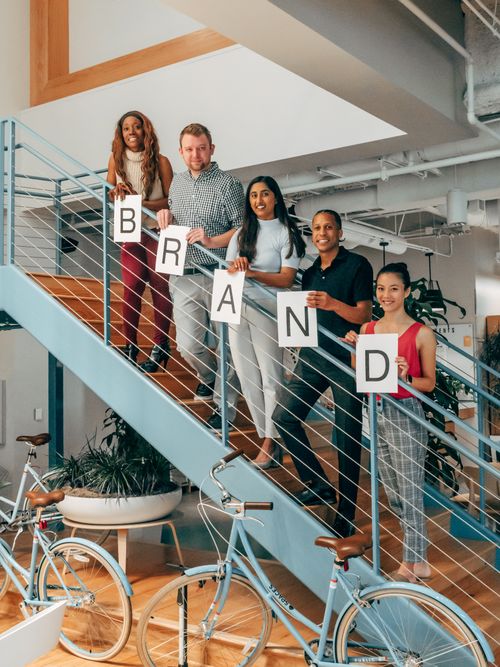 Free A diverse group of colleagues smiling and holding a 'Brand' sign on indoor office stairs. Stock Photo