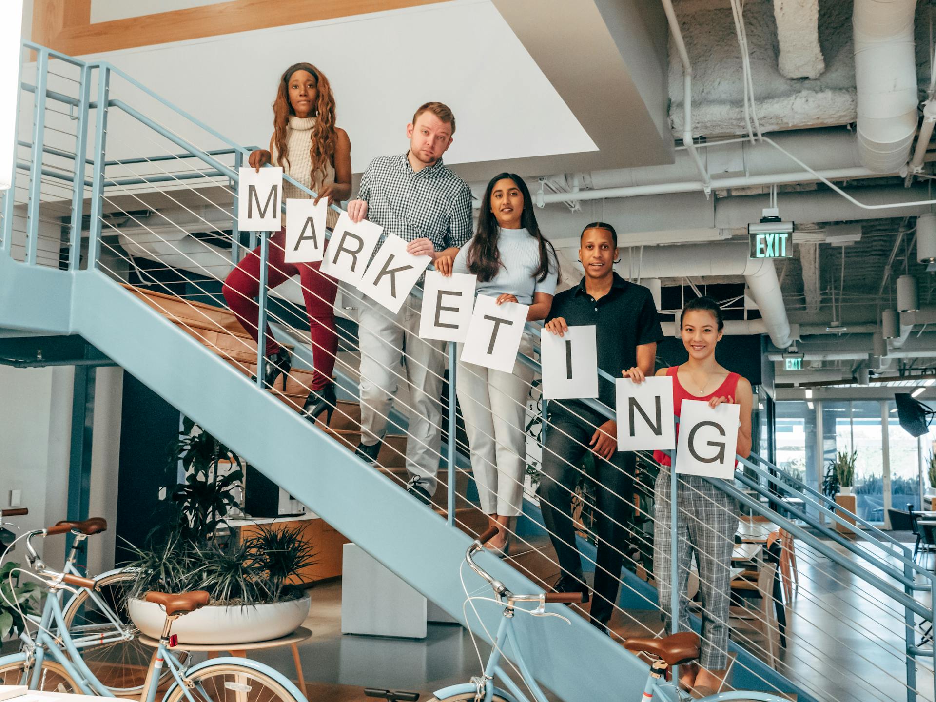 A diverse team of coworkers in an office holding a marketing sign on a staircase, smiling and happy.