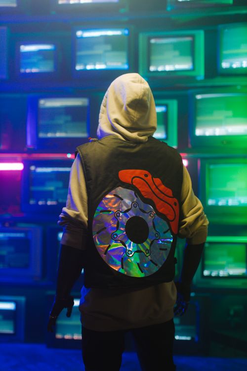 Free Person wearing a trendy hooded outfit stands before a wall of retro TVs, showcasing unique streetwear fashion. Stock Photo