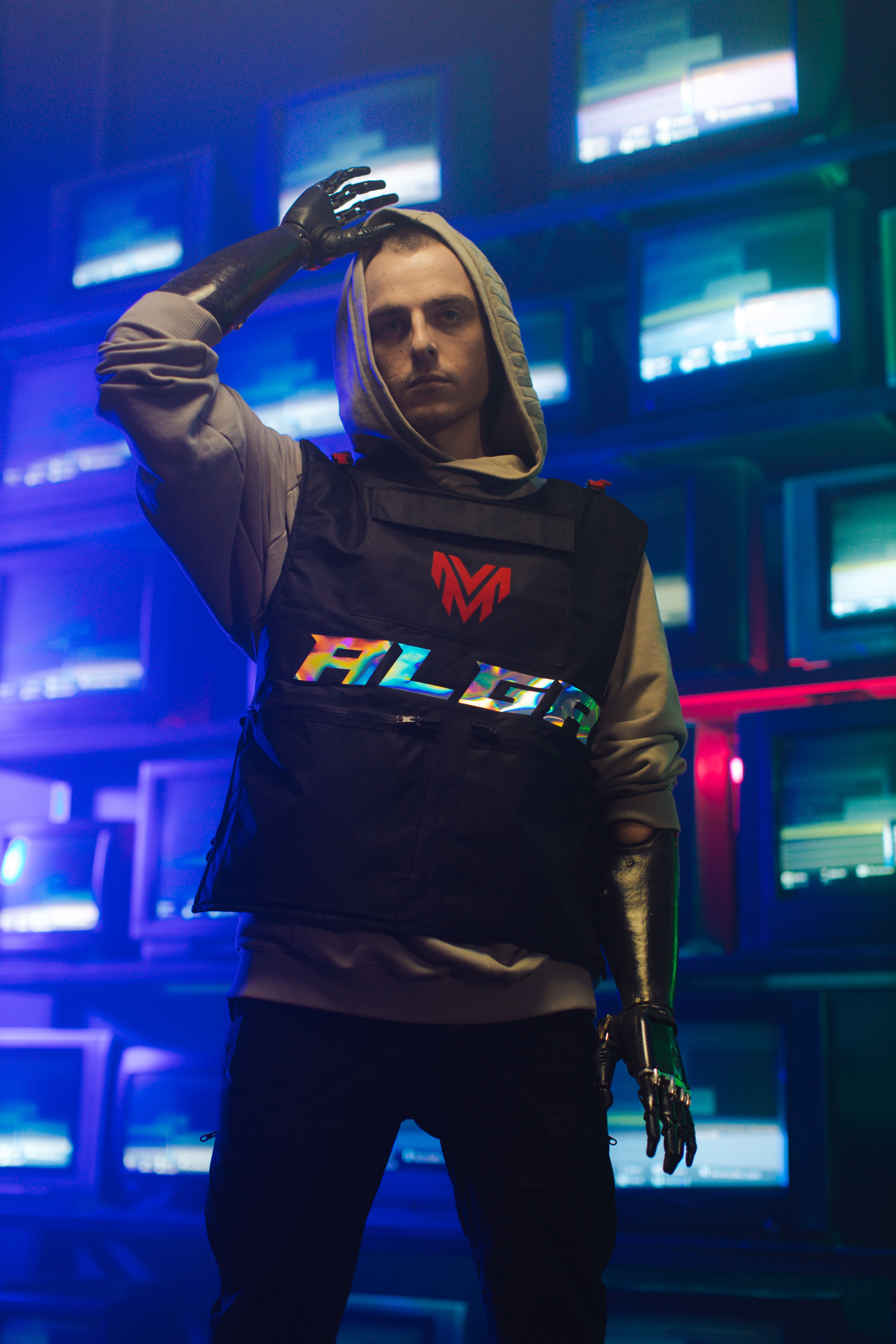 Model wearing a futuristic outfit with a prosthetic arm against a backdrop of neon-lit screens.