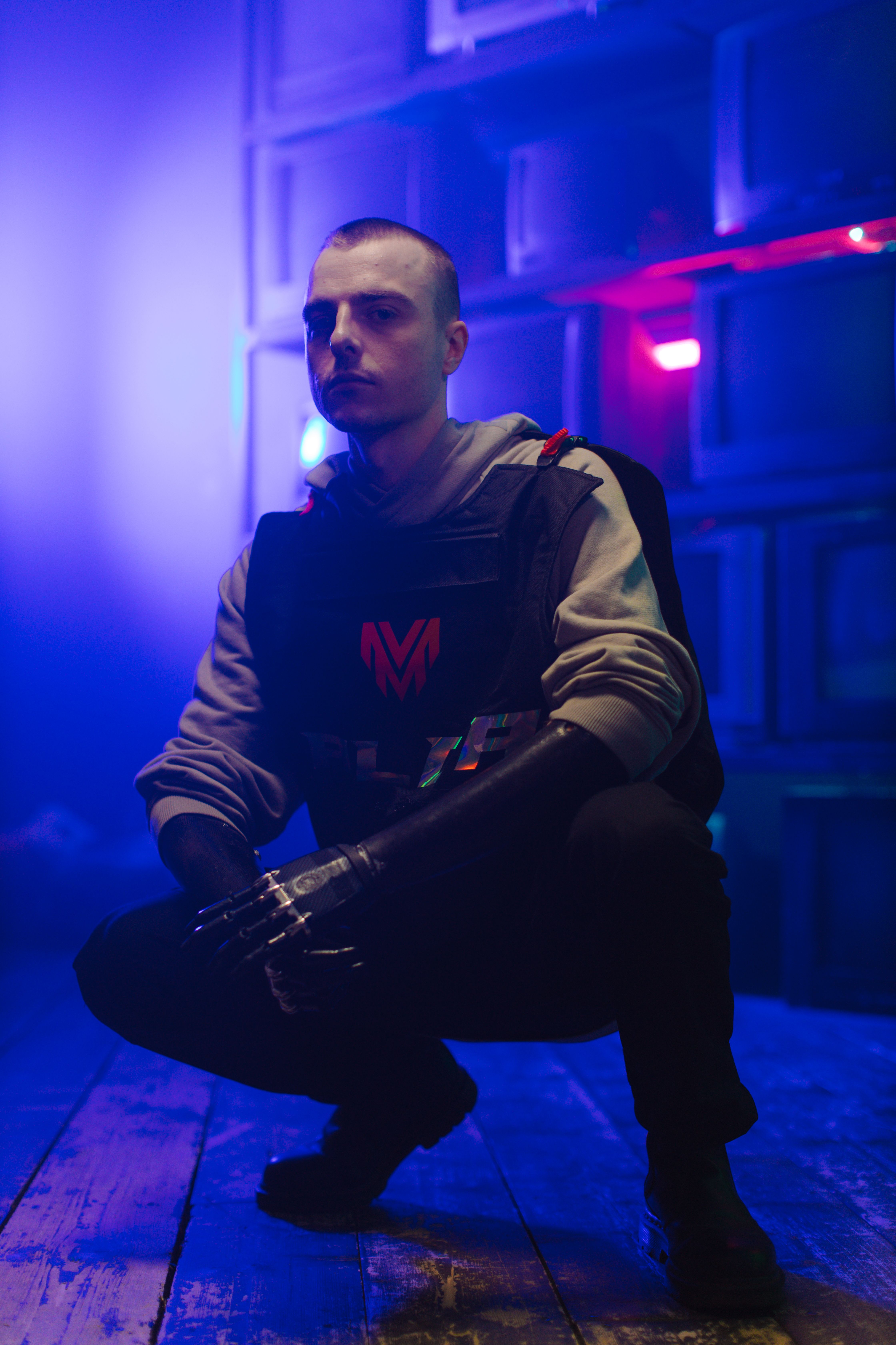Man in cyberpunk attire squatting under vibrant neon lights indoors.