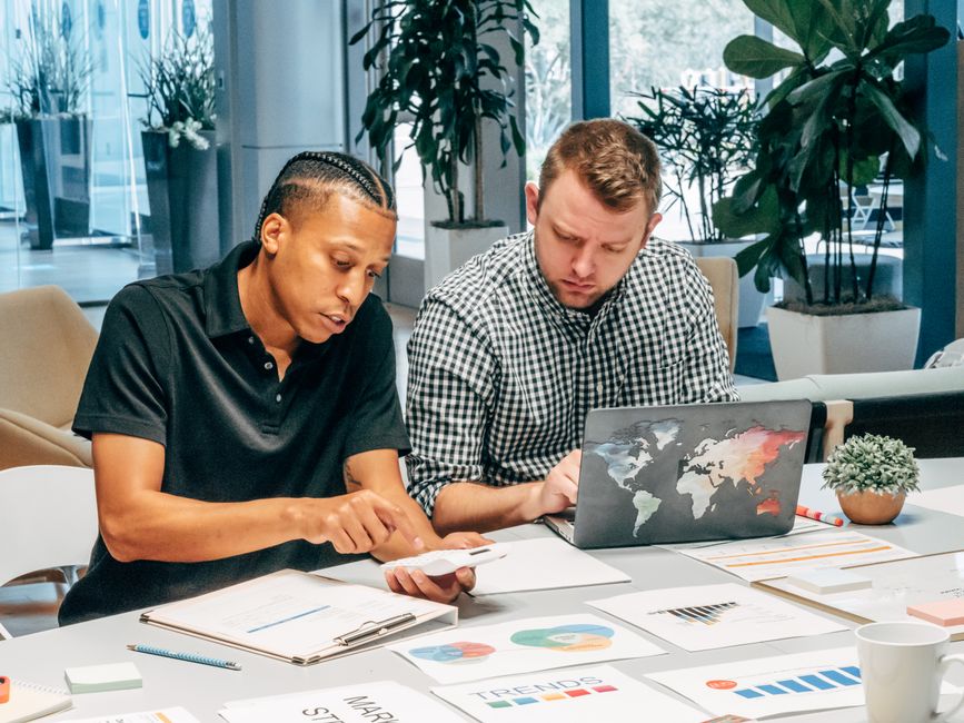 Two colleagues working together on a laptop and documents in an office setting.