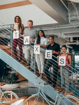 A group of diverse colleagues holding 'TAXES' sign while standing on a modern office staircase.
