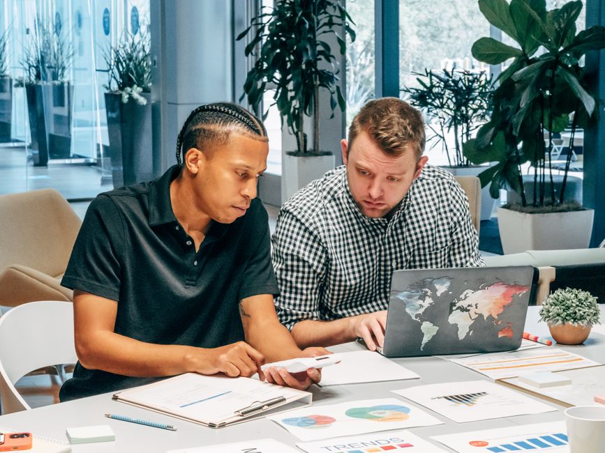 Two men working together in a modern office with laptops and documents, discussing strategies. Two men working together in a modern office with laptops and documents, discussing strategies.