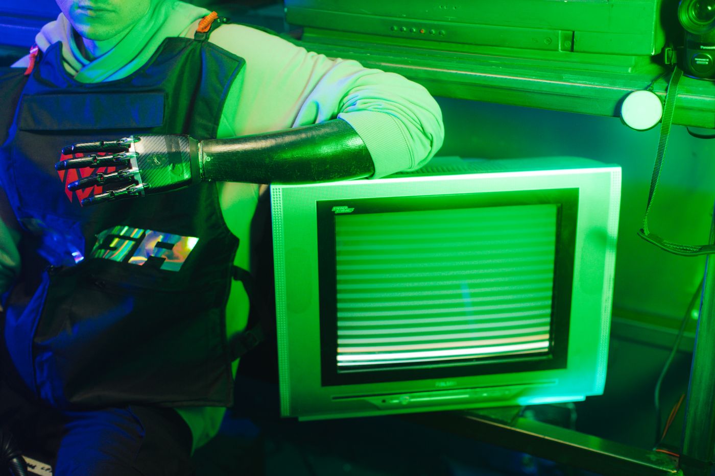 A close-up of a person with a bionic arm leaning on a CRT TV under green lighting.