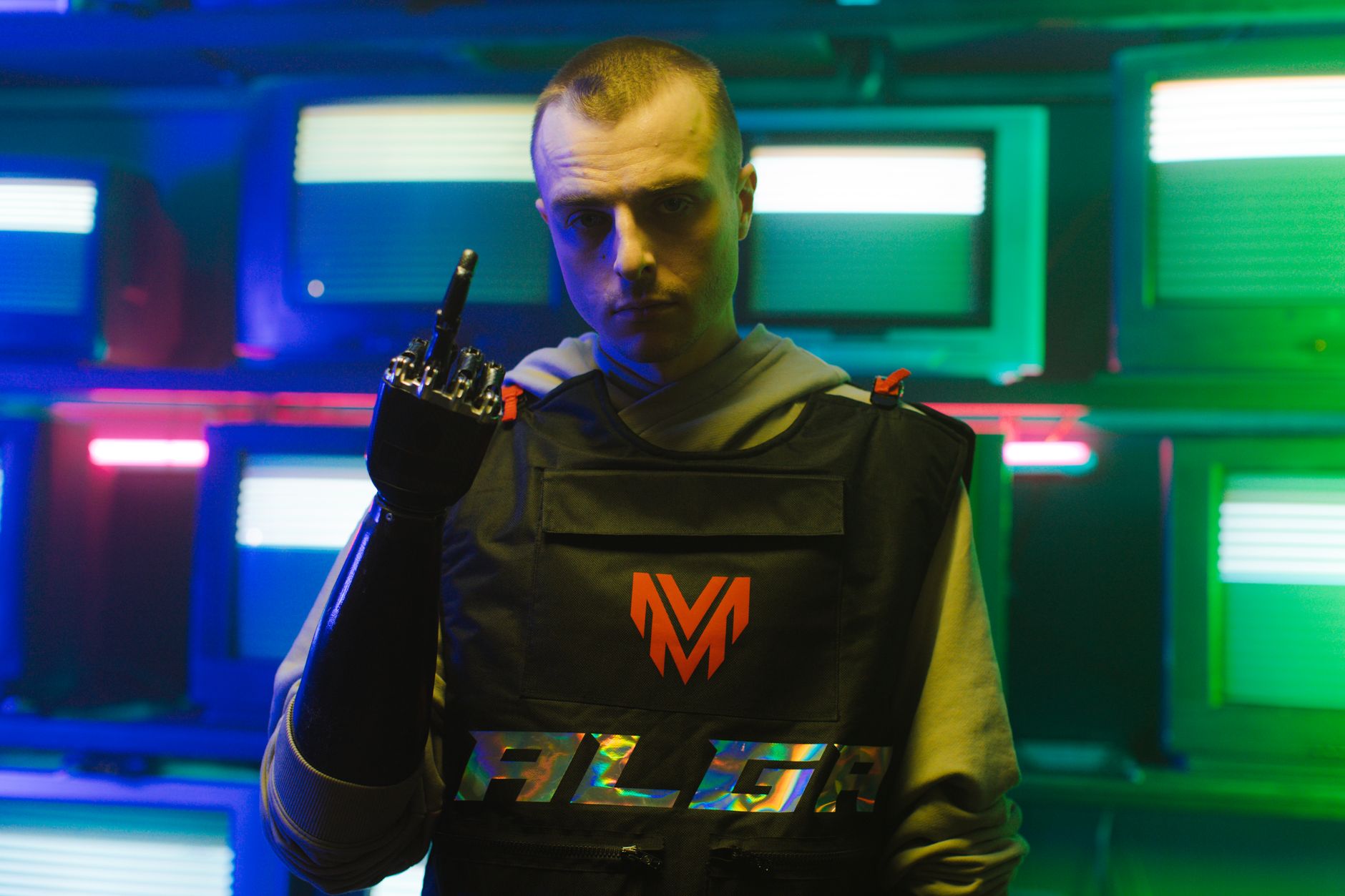 A man with a bionic arm stands confidently in a neon-lit room full of vintage TVs.