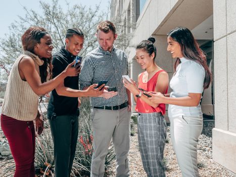 A diverse group of colleagues smiling and engaging with smartphones outside an office building.