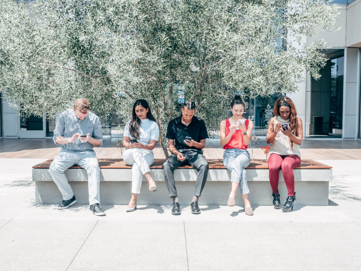 Group of friends sitting on a bench outdoors, each using a smartphone