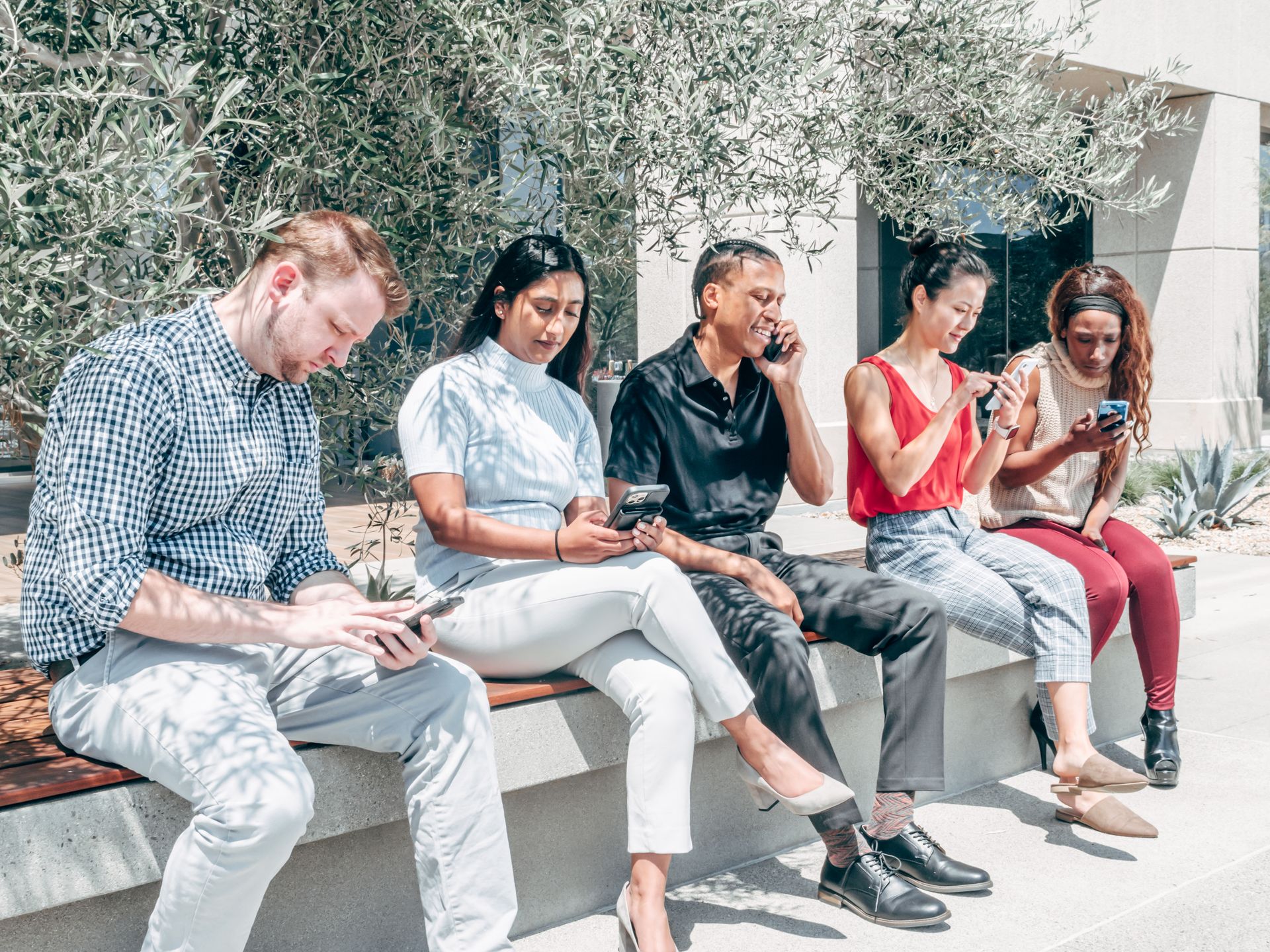 A diverse group of adults using smartphones while sitting on an outdoor bench.