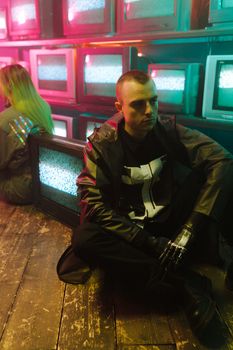 Stylish man with prosthetic arm in neon-lit room surrounded by vintage TVs.