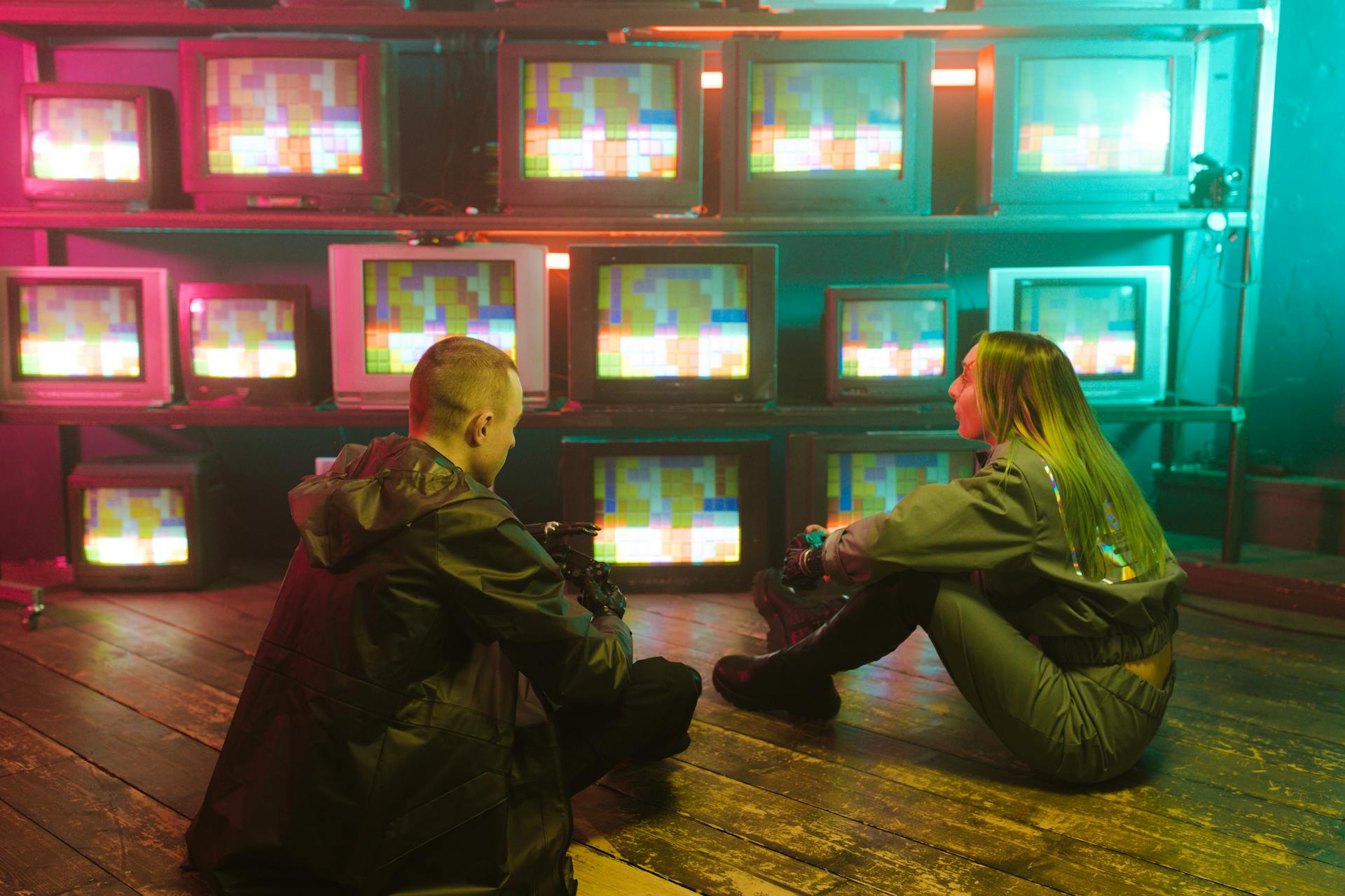 Two people sitting in a retro setting surrounded by neon-lit CRT TVs displaying pixel art.