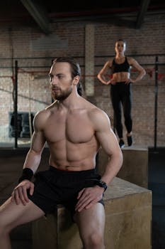 Muscular man resting on a plyo box indoors, fitness motivation in a gym setting.