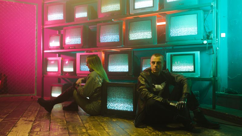 Two individuals in a cyberpunk setting with neon lighting and stacked television screens.