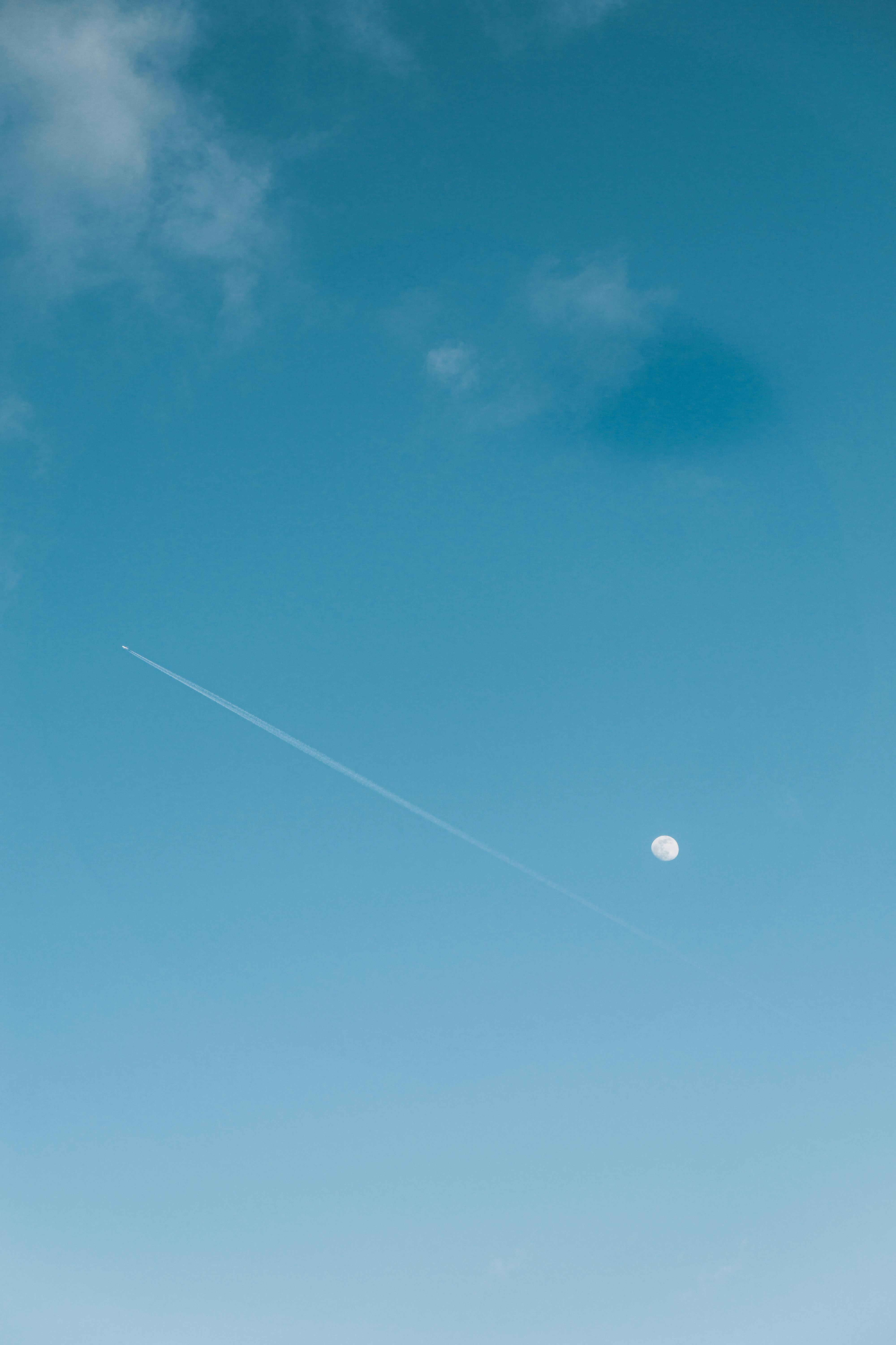 A serene sky photograph featuring a moon and airplane contrail against a clear blue backdrop.