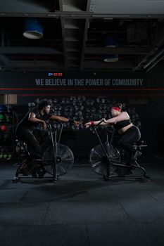 Fitness-focused diverse duo connecting on exercise bikes in a gym.