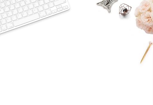 A top-down view of a white desk setup with floral decor, featuring a keyboard, pen, and flowers.