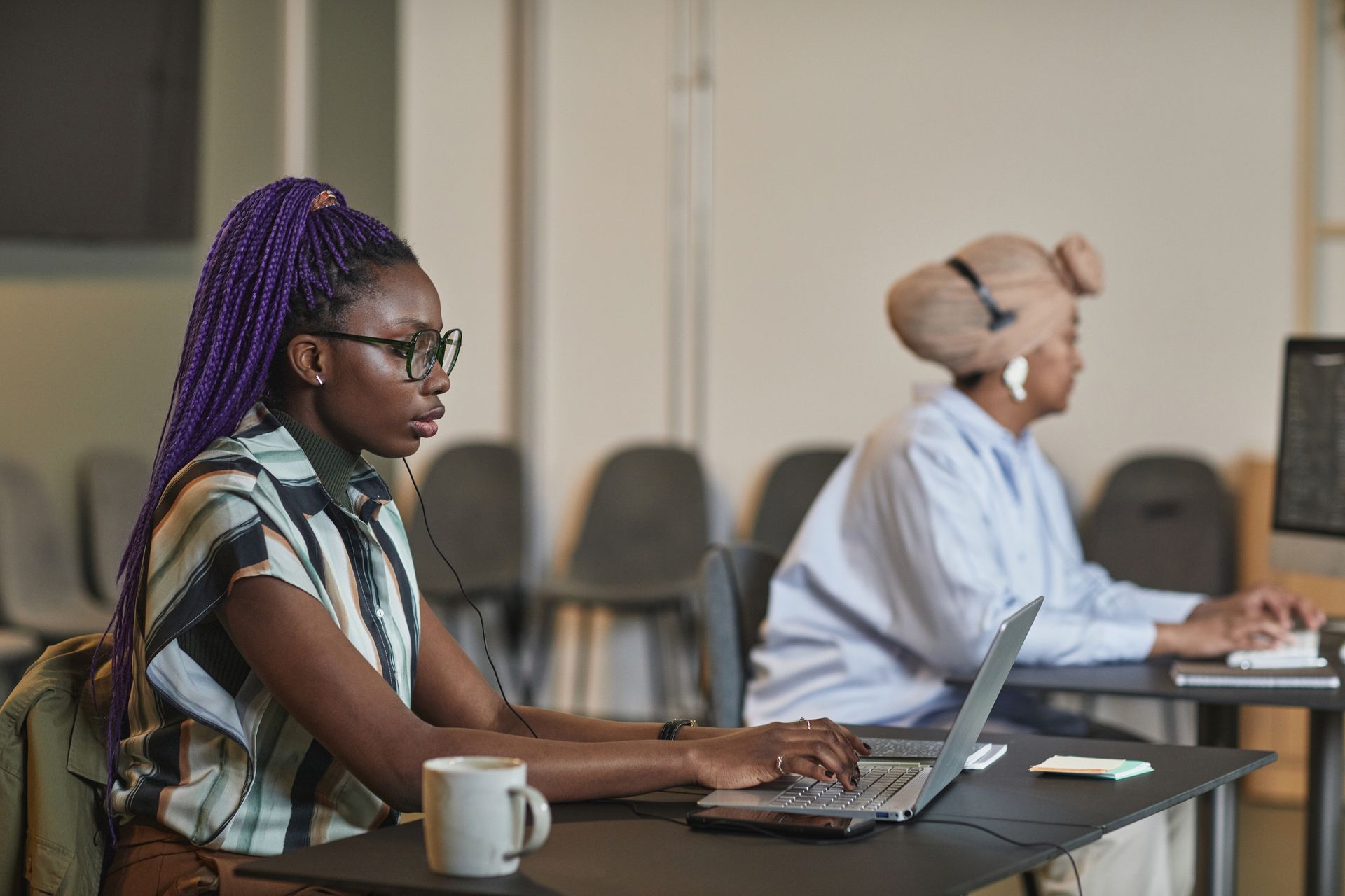 Two Black women working diligently at laptops in a modern office setting.