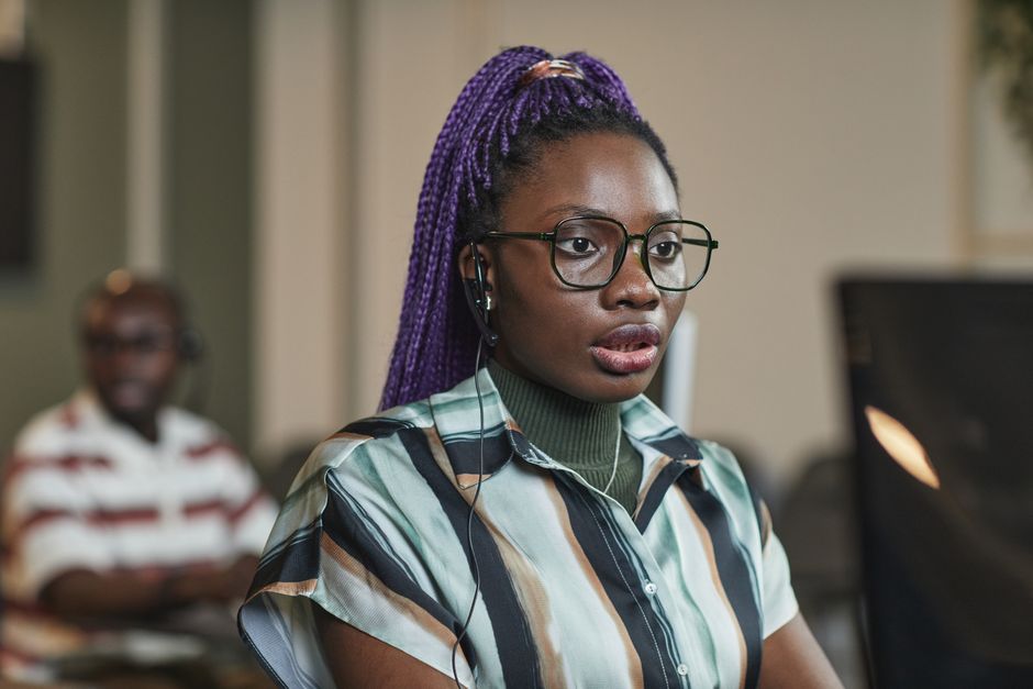 African-American woman with glasses and headset working in a modern office setting, showcasing concentration.