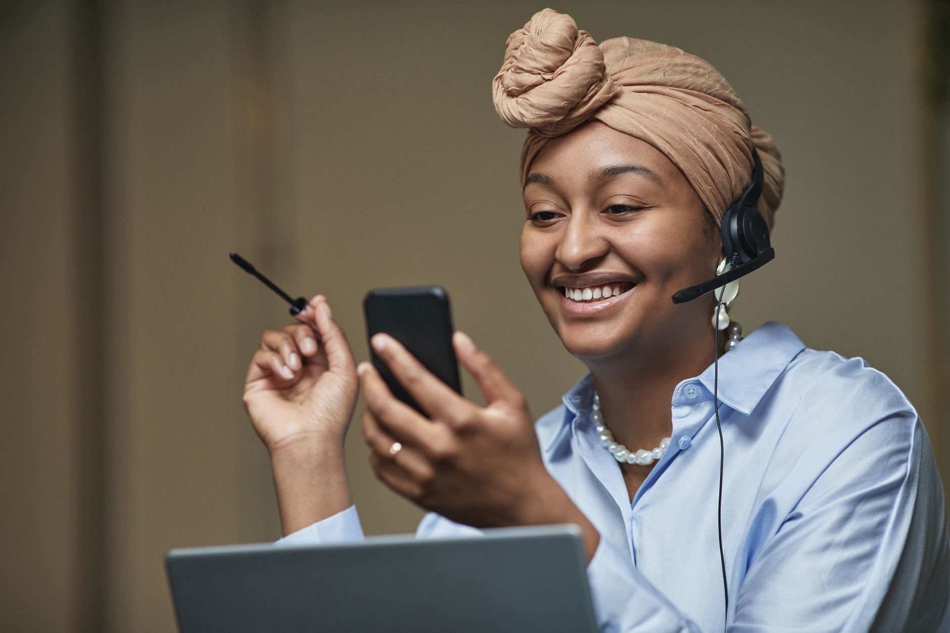 How to Fix Facebook App Freezing on Your Phone (Android and iPhone) A cheerful woman in a headwrap and headset multitasking between smartphone and laptop in an indoor office setting.
