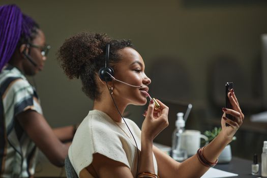 Two women multitasking in a call center, using headsets while applying makeup. Modern office life.