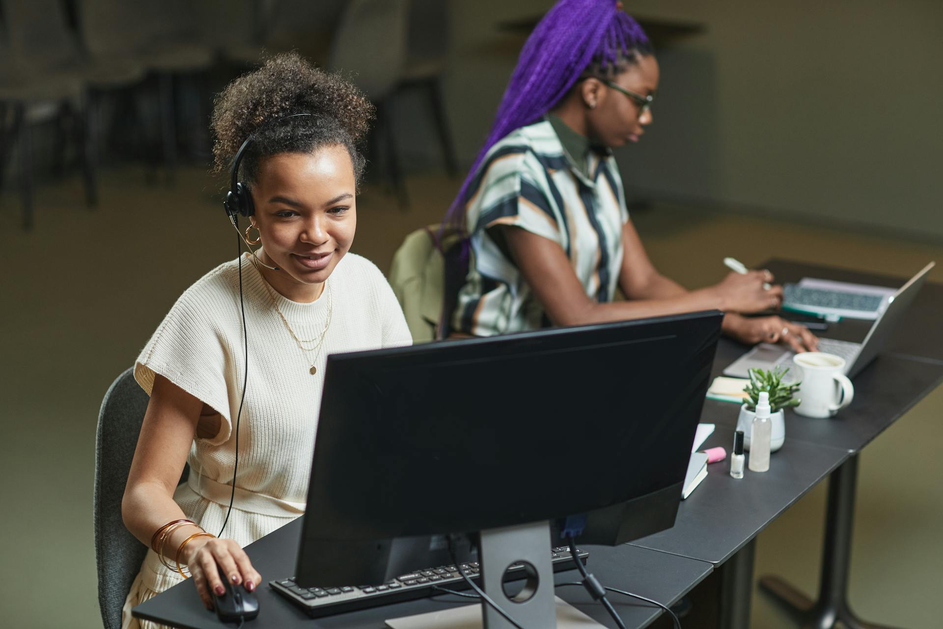 Two women working with computers and headsets in a modern office setting, showcasing teamwork and technology.