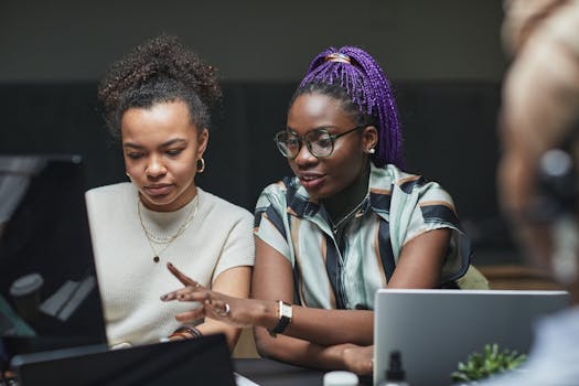 Two women working together on a project using laptops, showcasing teamwork and collaboration in a modern office setting.