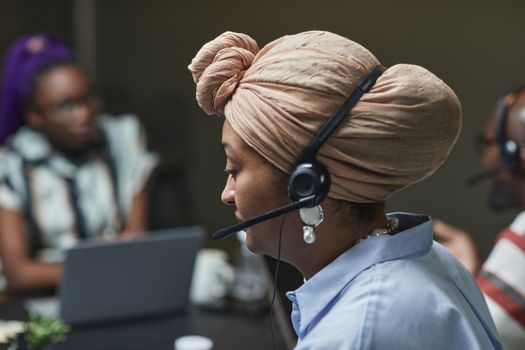 Side profile of a woman in a headscarf working in a call center, wearing a headset and microphone.