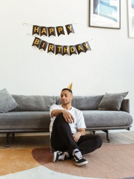 A young man wearing a party hat celebrates a birthday alone indoors with minimal decor.