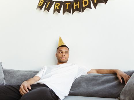 Young man in a white t-shirt sitting on a sofa with a party hat under a birthday banner.