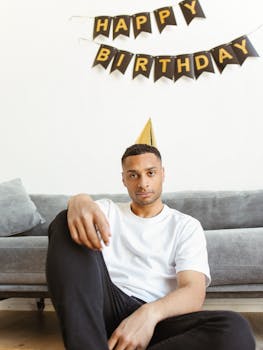 Man sitting on sofa, wearing party hat with happy birthday banner in background.