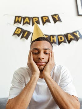 Man wearing a party hat looking thoughtful indoors with a birthday banner background