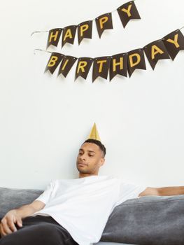 Reflective man in party hat under a 'Happy Birthday' banner, sitting on a sofa.