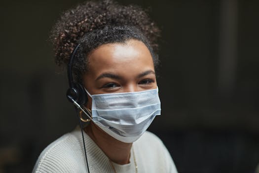 Portrait of a call center agent with curly hair and mask, wearing a headset indoors.