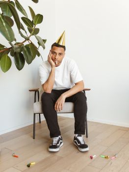 Young man in party hat sitting indoors looking pensive, surrounded by party favors.