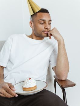 A man in a party hat looks pensive with a cupcake on his lap indoors.
