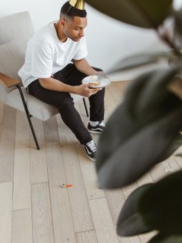 Adult man in casual attire with a party hat enjoying a piece of cake indoors.