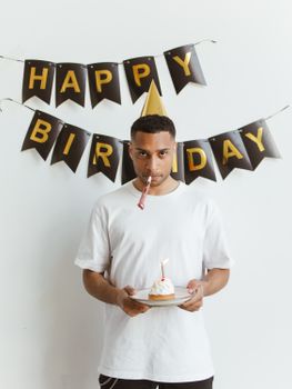 A young man in a party hat holds a cupcake under a happy birthday banner.